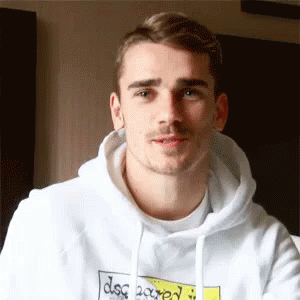 A Young Man Wearing A White Hoodie With A Yellow Sticker On It Is Looking At The Camera . GIF