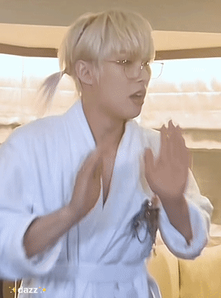 A Young Man Wearing A White Robe And Glasses Is Standing In A Room . GIF