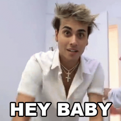 A Young Man Wearing A White Shirt And A Cross Necklace Is Standing In A Room And Saying `` Hey Baby '' . GIF