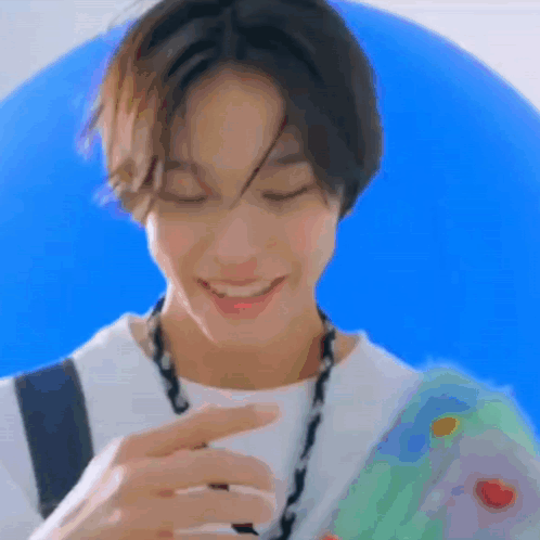 A Young Man Wearing A White Shirt And A Necklace Is Smiling While Holding A Blue Balloon . GIF