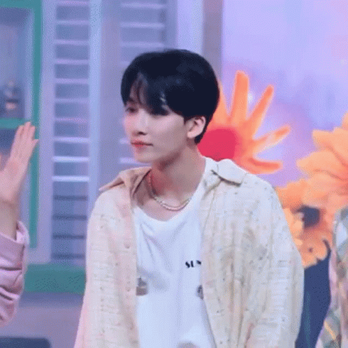 A Young Man Wearing A White Shirt And A Pearl Necklace Is Standing In Front Of A Bunch Of Flowers . GIF