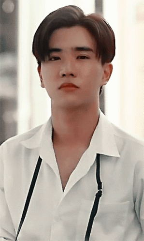 A Young Man Wearing A White Shirt With A Black Strap Around His Neck Looks At The Camera GIF
