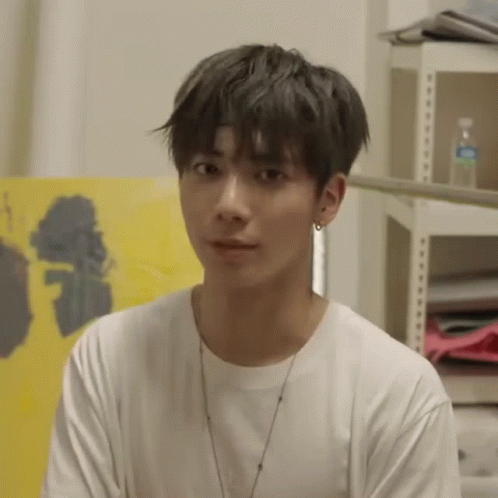 A Young Man Wearing A White T-Shirt And A Necklace Is Sitting In Front Of A Painting . GIF