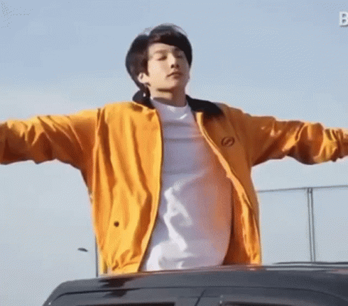 A Young Man Wearing A Yellow Jacket And A White Shirt Is Standing In Front Of A Car With His Arms Outstretched . GIF