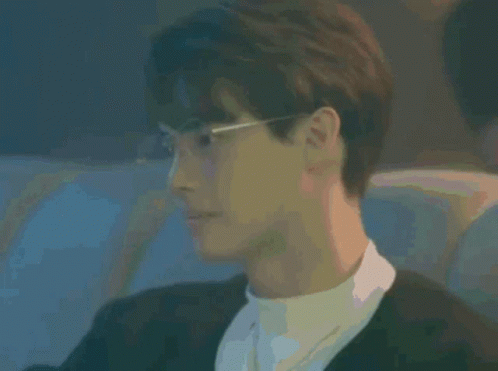 A Young Man Wearing Glasses And A Black Jacket Is Sitting In A Dark Room . GIF