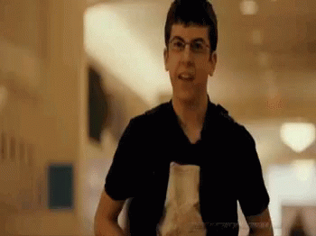 A Young Man Wearing Glasses And A Black Shirt Is Holding A Piece Of Paper In A Hallway . GIF