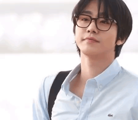 A Young Man Wearing Glasses And A Blue Shirt Is Standing In Front Of A Body Of Water . GIF