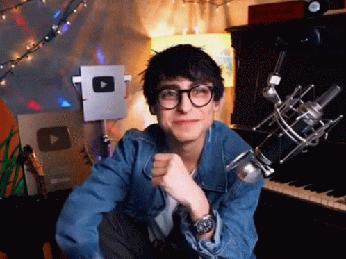 A Young Man Wearing Glasses And A Denim Jacket Is Sitting In Front Of A Microphone And A Piano . GIF