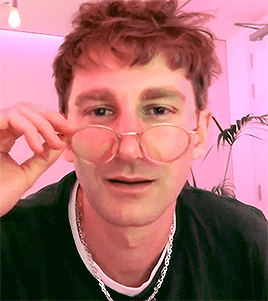 A Young Man Wearing Glasses And A Necklace Is Looking At The Camera . GIF