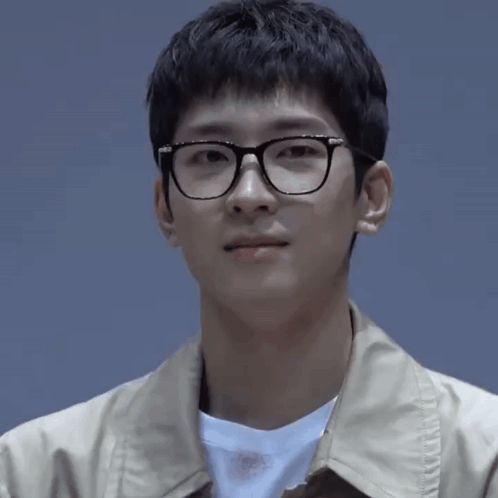 A Young Man Wearing Glasses And A Tan Jacket Is Looking At The Camera GIF