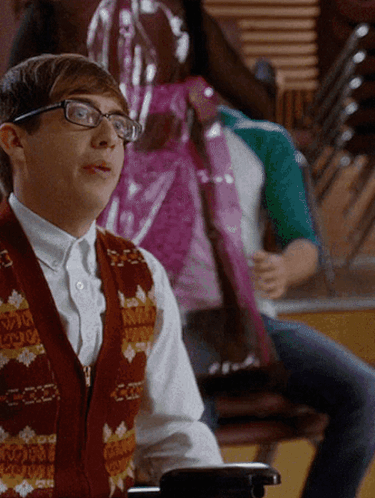 A Young Man Wearing Glasses And A Vest That Says ' A ' On It Sits In A Chair GIF