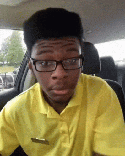 A Young Man Wearing Glasses And A Yellow Shirt Is Sitting In The Driver 'S Seat Of A Car GIF