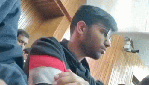 A Young Man Wearing Glasses Is Sitting At A Table In A Room . GIF
