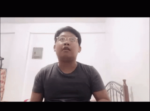 A Young Man Wearing Glasses Is Sitting In Front Of A Bed . GIF
