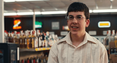 A Young Man Wearing Glasses Stands In Front Of A Shelf Of Liquor Bottles GIF