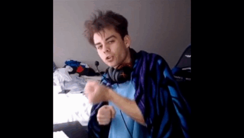 A Young Man Wearing Headphones And A Blue And Purple Blanket Is Sitting On A Bed . GIF