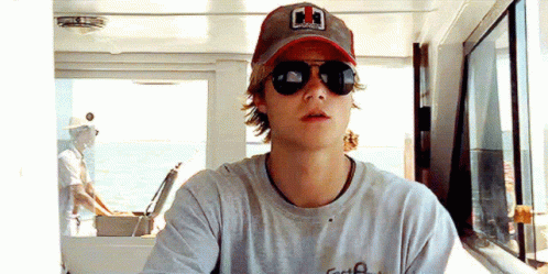 A Young Man Wearing Sunglasses And A Baseball Cap Is Sitting On A Boat . GIF
