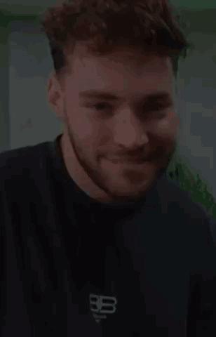 A Young Man With A Beard Is Smiling And Wearing A Black T-Shirt . GIF