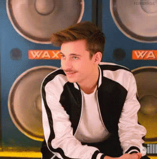 A Young Man With A Mustache Is Sitting In Front Of A Wall Of Speakers With The Word Wax On It . GIF