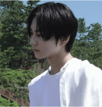 A Young Man With Black Hair And A White Shirt Is Standing In Front Of Trees GIF