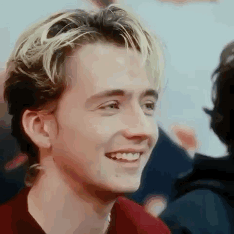 A Young Man With Blonde Hair And A Red Shirt Is Smiling And Looking At The Camera . GIF