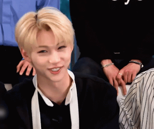 A Young Man With Blonde Hair And A White Scarf Around His Neck Is Smiling While Sitting In A Room With Other People . GIF