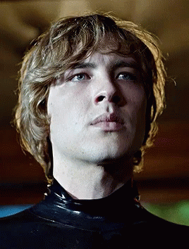A Young Man With Blonde Hair And Blue Eyes Is Wearing A Black Latex Bodysuit GIF
