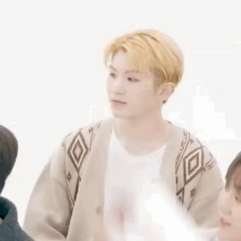 A Young Man With Blonde Hair Is Sitting In Front Of A White Wall With His Arm In The Air . GIF