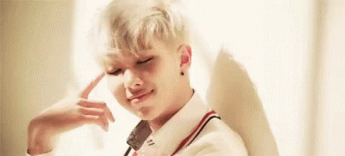 A Young Man With Blonde Hair Is Sitting In Front Of A Window With His Hand On His Head . GIF