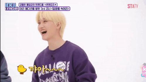 A Young Man With Blonde Hair Is Smiling And Holding A Duck In Front Of A Screen That Says Idol League GIF