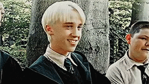 A Young Man With Blonde Hair Is Smiling While Standing Next To A Group Of Young Men . GIF
