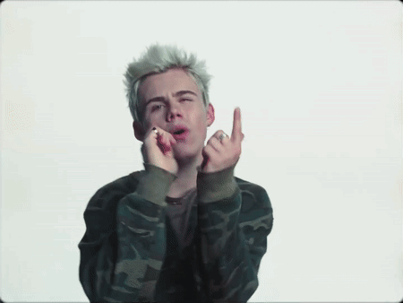 A Young Man With Blonde Hair Is Wearing A Camouflage Jacket And Giving The Middle Finger . GIF