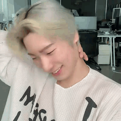 A Young Man With Blonde Hair Is Wearing A White Sweater With A Black Letter T On It . GIF