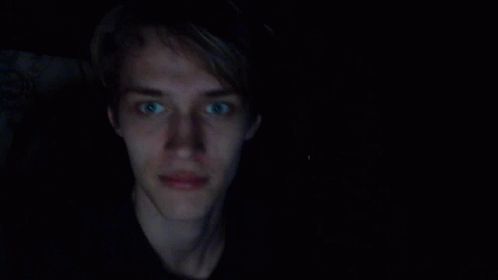 A Young Man With Blue Eyes Is Smiling In A Dark Room GIF