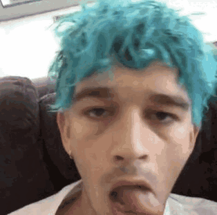 A Young Man With Blue Hair Is Sticking His Tongue Out While Sitting On A Couch . GIF