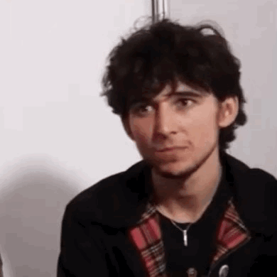 A Young Man With Curly Hair And A Beard Is Wearing A Plaid Shirt And A Black Jacket . GIF