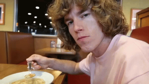 a young man with curly hair is sitting at a table with a plate of food and a fork in his hand .