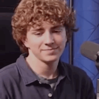 A Young Man With Curly Hair Is Sitting In Front Of A Microphone . GIF