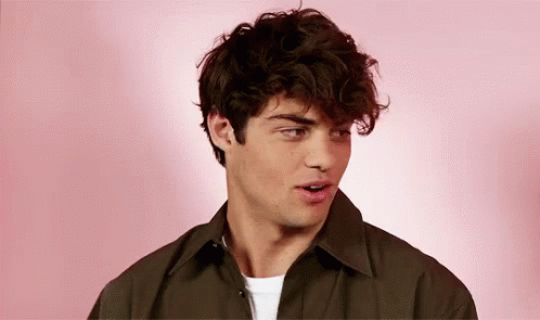 A Young Man With Curly Hair Is Standing In Front Of A Pink Background And Looking At The Camera . GIF