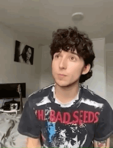 A Young Man With Curly Hair Is Wearing A Bad Seeds T-Shirt And Looking At The Camera . GIF