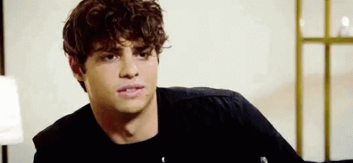 A Young Man With Curly Hair Is Wearing A Black Shirt And Sitting In A Room . GIF