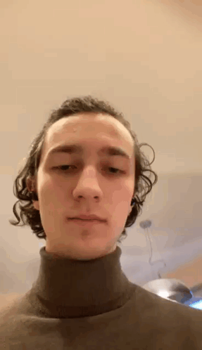 A Young Man With Curly Hair Is Wearing A Black Turtleneck Sweater And Looking At The Camera . GIF
