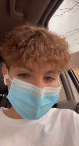 A Young Man With Curly Hair Is Wearing A Blue Face Mask While Sitting In A Car . GIF
