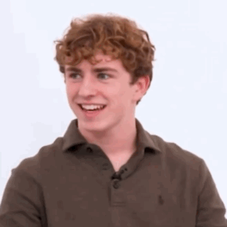A Young Man With Curly Hair Is Wearing A Brown Polo Shirt And Smiling . GIF