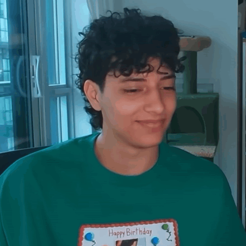 A Young Man With Curly Hair Is Wearing A Green Shirt And Holding A Happy Birthday Card . GIF