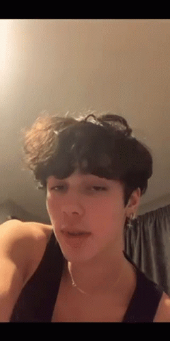 A Young Man With Curly Hair Is Wearing A Tank Top And Earrings . GIF
