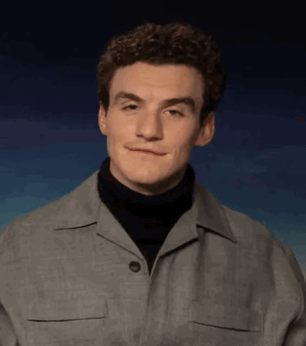 A Young Man With Curly Hair Is Wearing A Turtleneck And A Grey Jacket . GIF