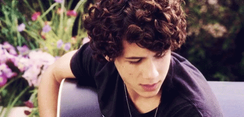 A Young Man With Curly Hair Wearing A Black Shirt And Necklace GIF