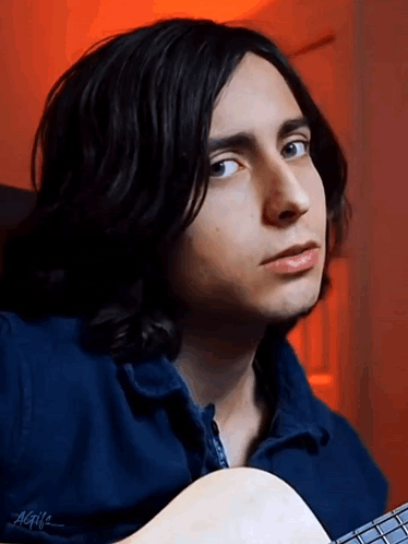 a young man with long black hair is holding a guitar and wearing a blue jacket