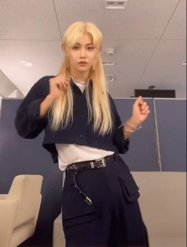 A Young Man With Long Blonde Hair Is Standing In A Room Wearing A Blue Jacket And Black Pants . GIF
